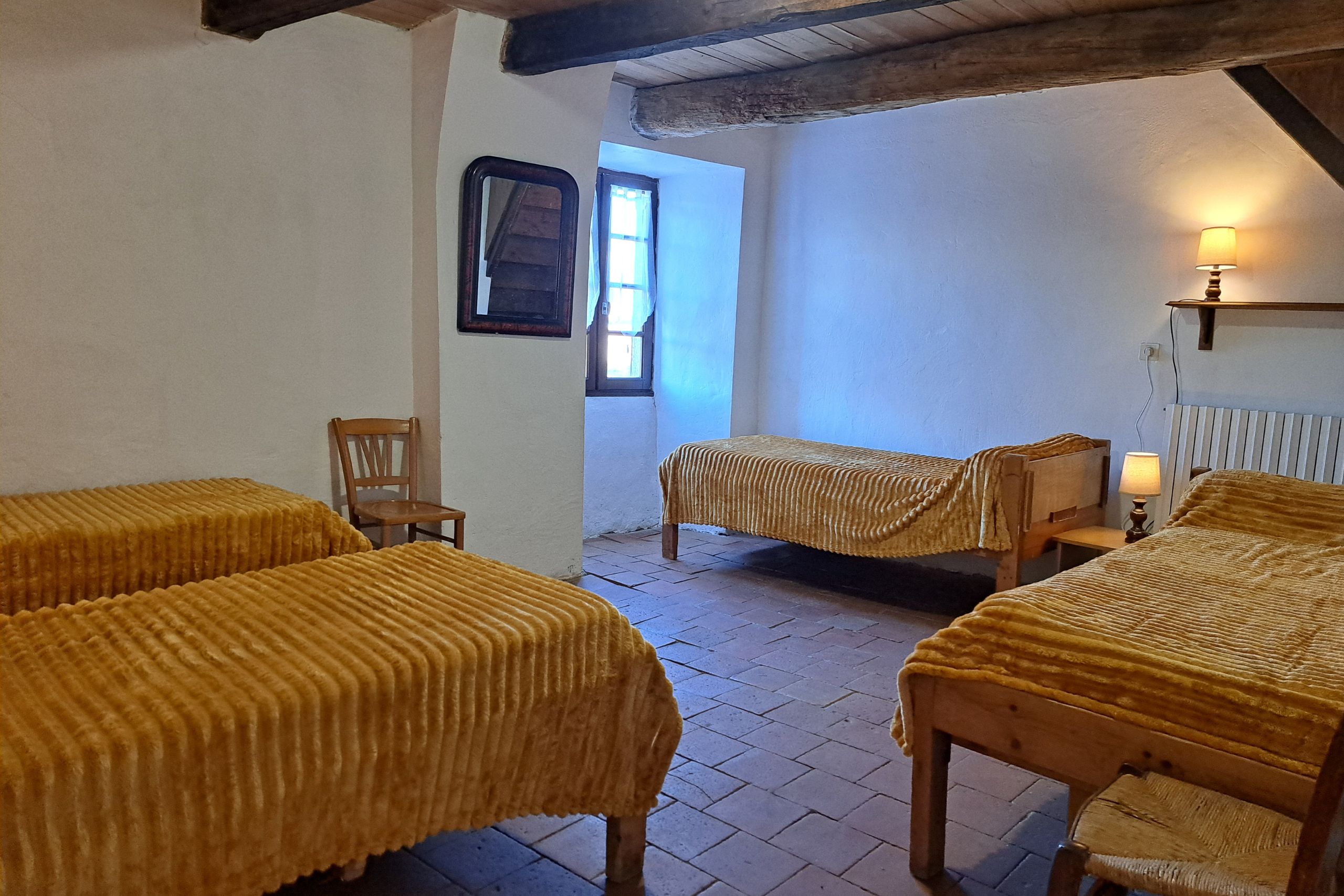 Chambre familiale avec quatre lits simples, sol en tomettes et poutres apparentes, gîte d’étape de la Tour du Viala‑du‑Pas‑de‑Jaux.