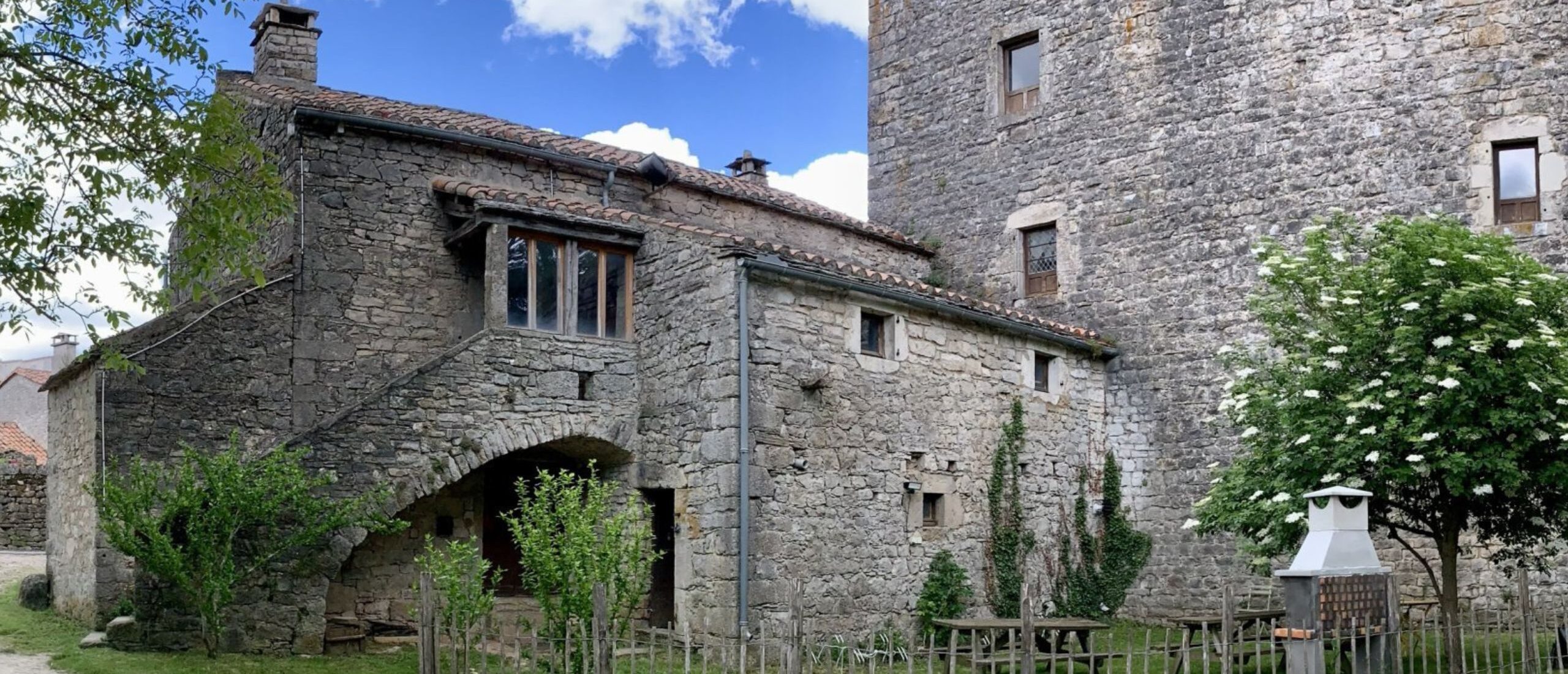 Vue extérieure du gîte en pierre adossé à la Tour du Viala-du-Pas-de-Jaux avec jardin et mobilier extérieur.