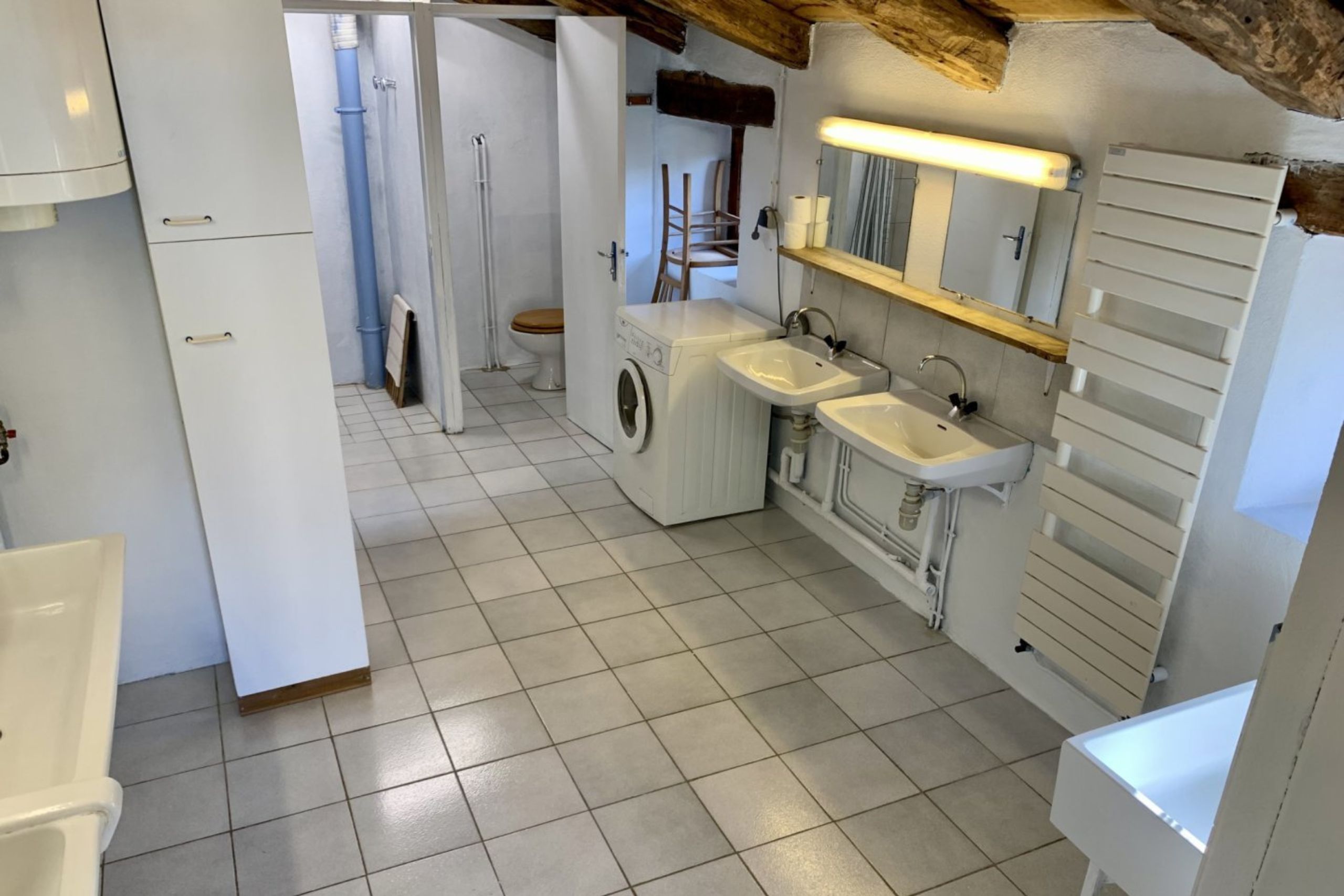 Salle de bain du gîte avec douches, lavabos, toilettes, machine à laver et poutres apparentes sous toiture.