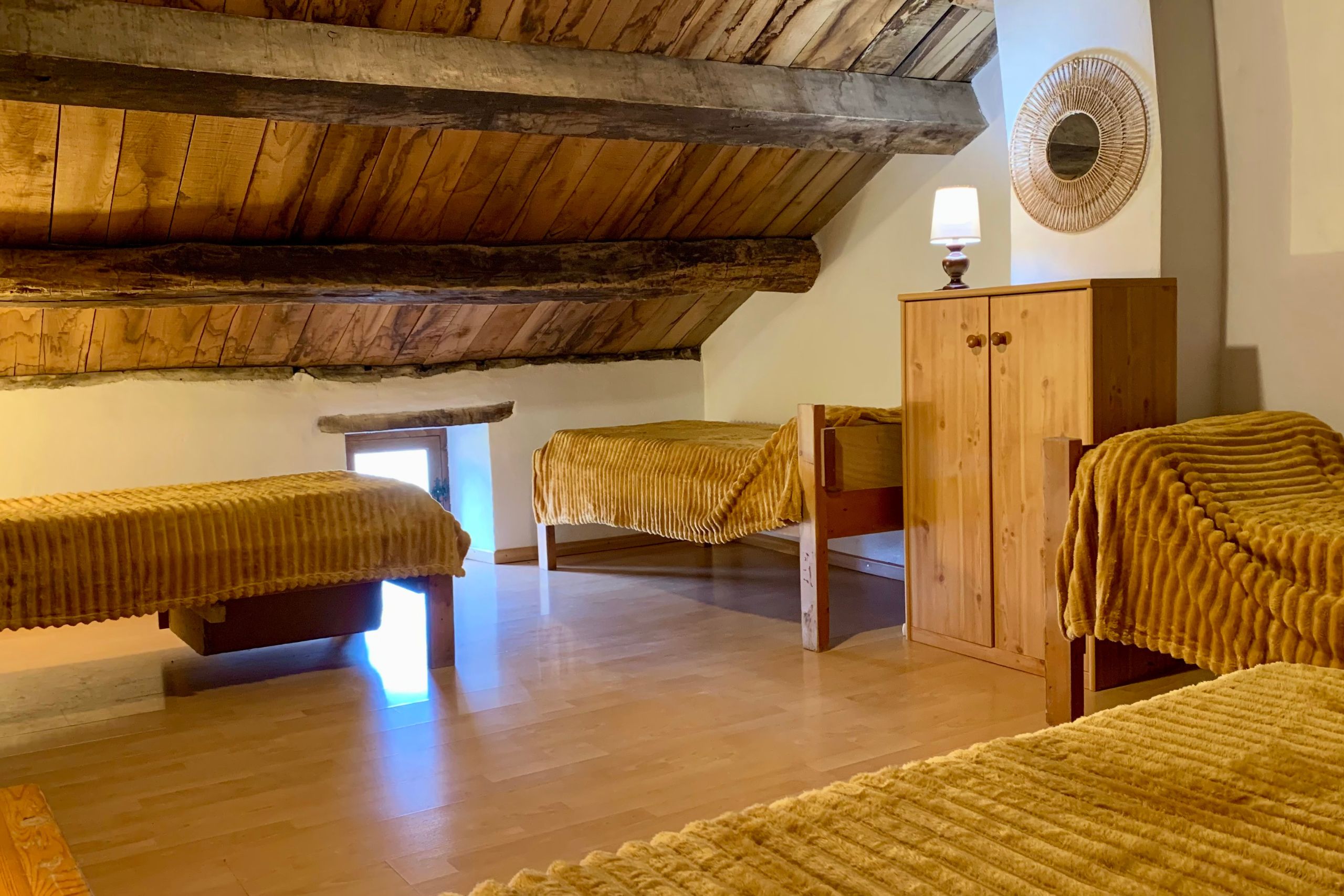 Chambre à l’étage du gîte d’étape de la Tour du Viala‑du‑Pas‑de‑Jaux avec 4 lits simples, couverture ocre et charpente apparente en bois.