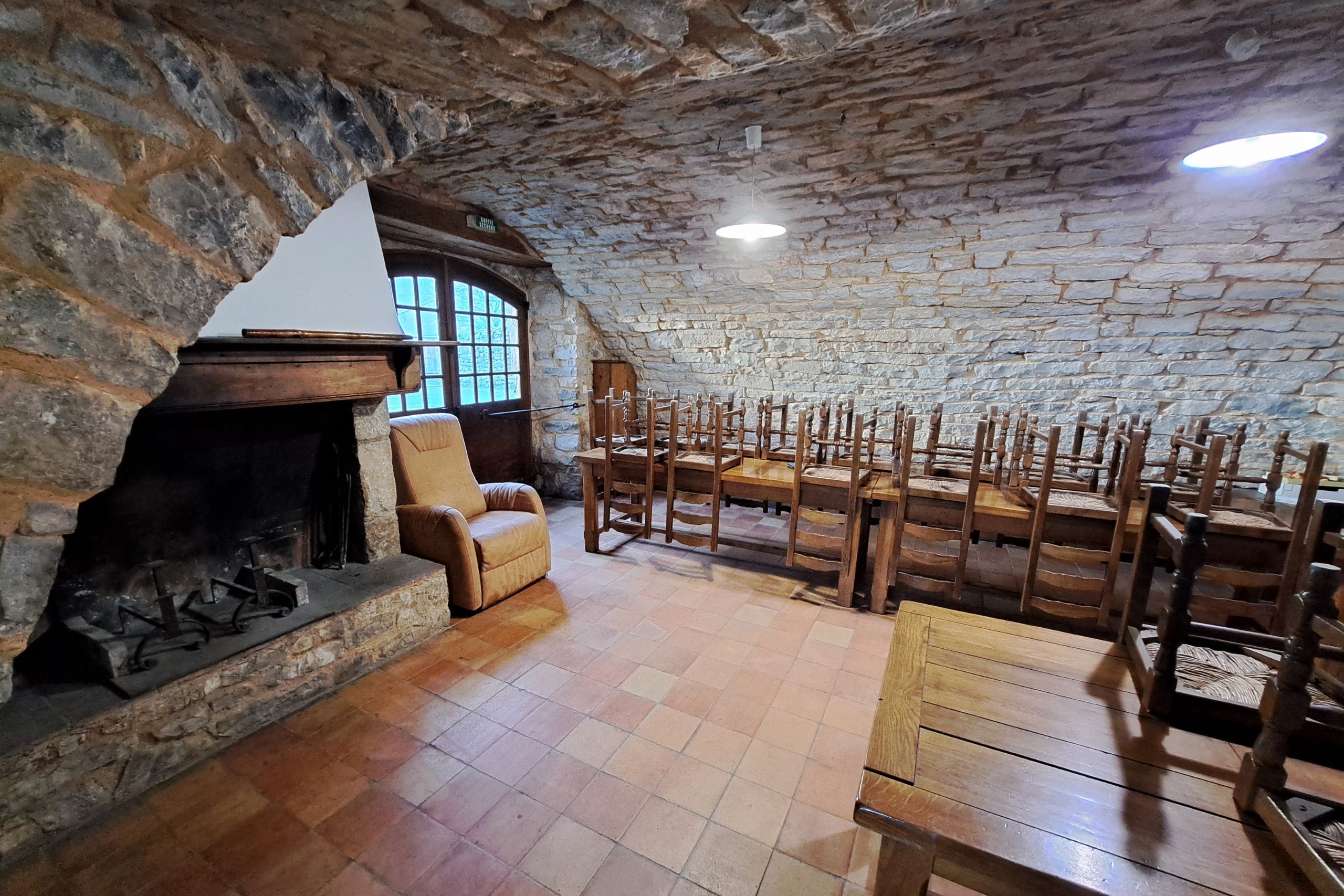 Salle commune avec cheminée en pierre, fauteuil et tables en bois dans le gîte d’étape de la Tour du Viala‑du‑Pas‑de‑Jaux.